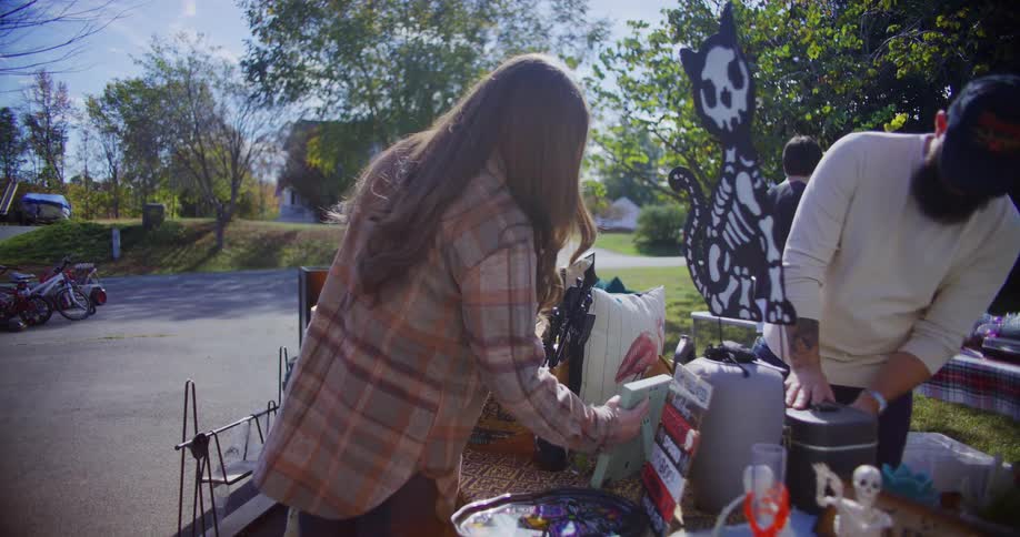 A woman browses through items at a yard sale while a man stands behind the table and a child approaches