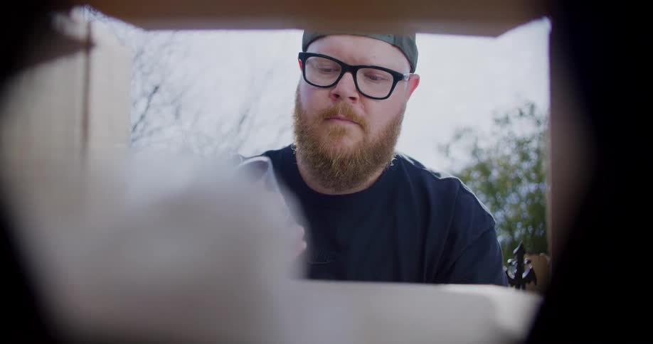 Seen from inside a box a bearded man opens the package removing and unwrapping two shot glasses