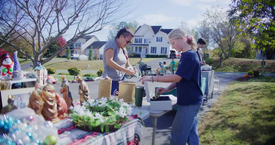 A woman is selling items including holiday decorations and a wreath to a young girl who is handling cash at a table during a busy outdoor yard sale in a suburban neighborhood