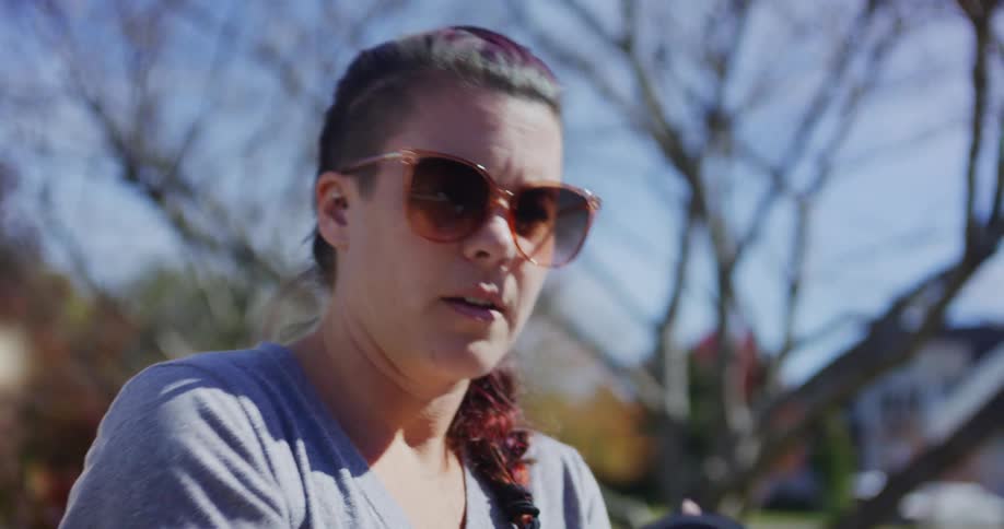 A woman picks up a digital camera from a desk outdoors at midday wearing sunglasses she raises it to try taking a photograph under the bright sun