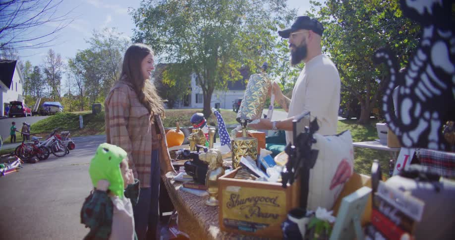A man wearing glasses a baseball hat and facial hair sorts through items at a garage sale he hands one to the seller for a price while a woman examines the item nearby beside her a child plays with a hat under the bright midday sun