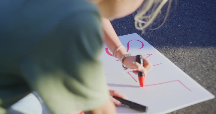 The video shows a closeup overtheshoulder view of a person with blonde hair wearing a green top they are crouched down outdoors drawing with a red marker on a white piece of poster board or similar material resting on the ground