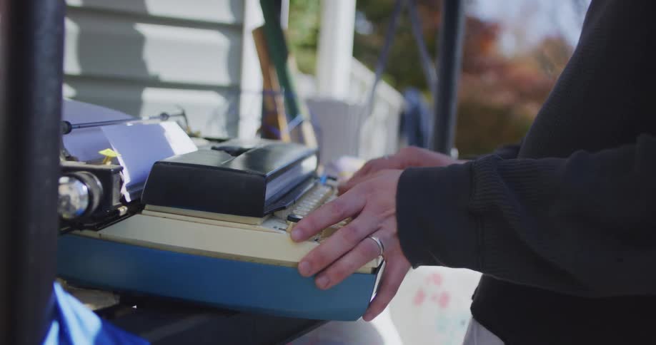 Young adult man pressing keys and testing a typewriter on a shelf next to a house outdoors during day time