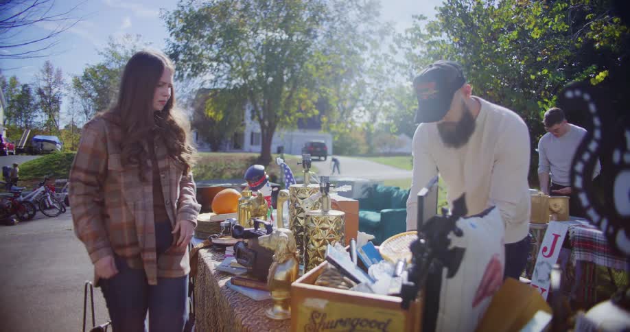 A woman in a plaid shirt stands in a driveway looking at a table piled high with various items for sale including several goldcolored decorative objects a bearded man in a white sweater and a baseball cap is standing across the table from her momenta