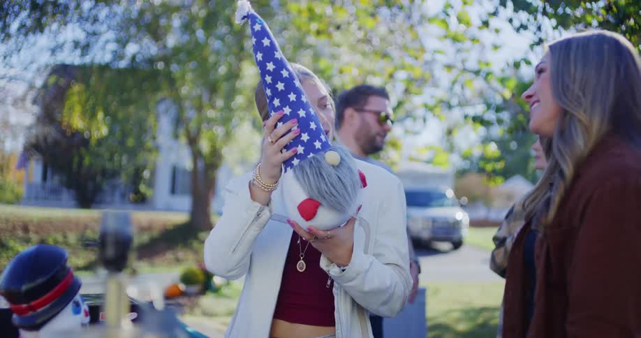 A young woman in a white jacket holds up a gnome decoration and smiles while talking to another woman at an outdoor flea market on a sunny day
