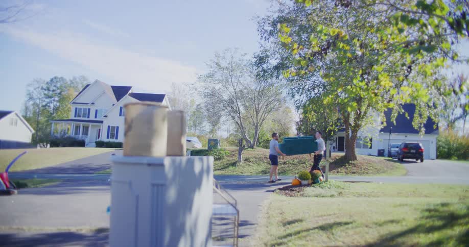 A wide shot shows two men carrying a large green plastic storage bin across a suburban lawn on a sunny day with houses and autumn trees in the background