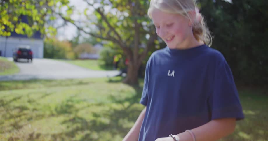 A young girl in a dark blue shirt and denim shorts counts money and puts it into her pocket then smiles while standing outside near a table and a sunny suburban street