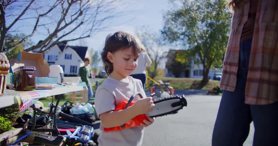 A young boy in a white shirt plays with an orange toy chainsaw next to a woman in a plaid jacket near a table of items at an outdoor yard sale on a bright sunny day