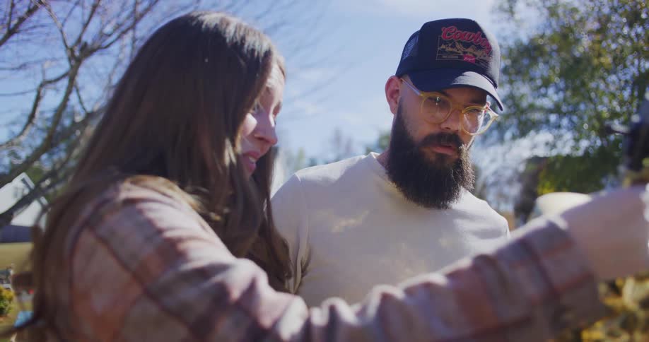 A woman stands outdoors holding an item while talking to a bearded man the man wearing a hat and glasses listens attentively as they engage in conversation