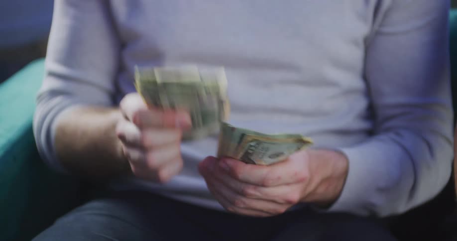 An adult male is holding and counting a stack of cash bills then hands over some money to a person next to him