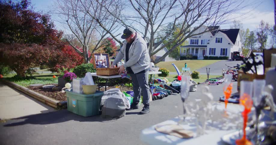 A man and a woman stand by a table full of merchandise as the man shows the woman a woven picnic basket during an outdoor garage sale on a sunny suburban driveway