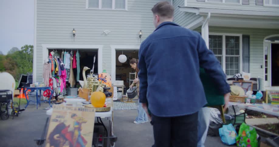 A woman in a plaid shirt directs two men carrying a green couch across a driveway full of items for a yard sale outside a suburban home