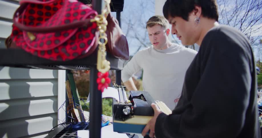 Two young men stand at a garage sale under the midday sun inspecting a typewriter placed on a shelf they examine it closely while engaged in discussion curious about its condition and value
