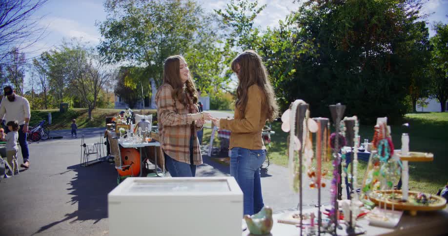 Two women shop outdoors at a garage sale on a sunny day browsing through items together around them other people and children move about adding to the lively atmosphere of the event