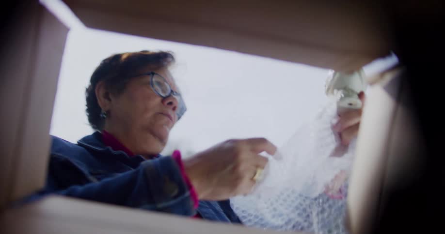 An older woman reaches into a box and unwraps a white ceramic bird figurine