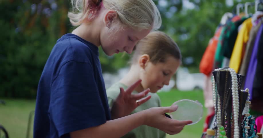 Two young girls browse items at an outdoor yard sale where one girl picks up and curiously examines a small round compact mirror as her friend looks on