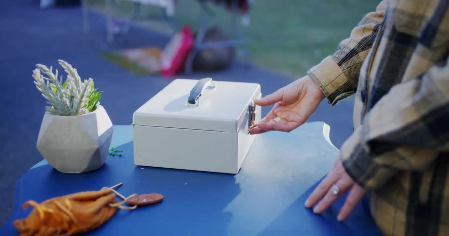 A cashier accepts payment from a customer during a garage sale at midday she places the money into the cash register while the customer purchases a potted plant