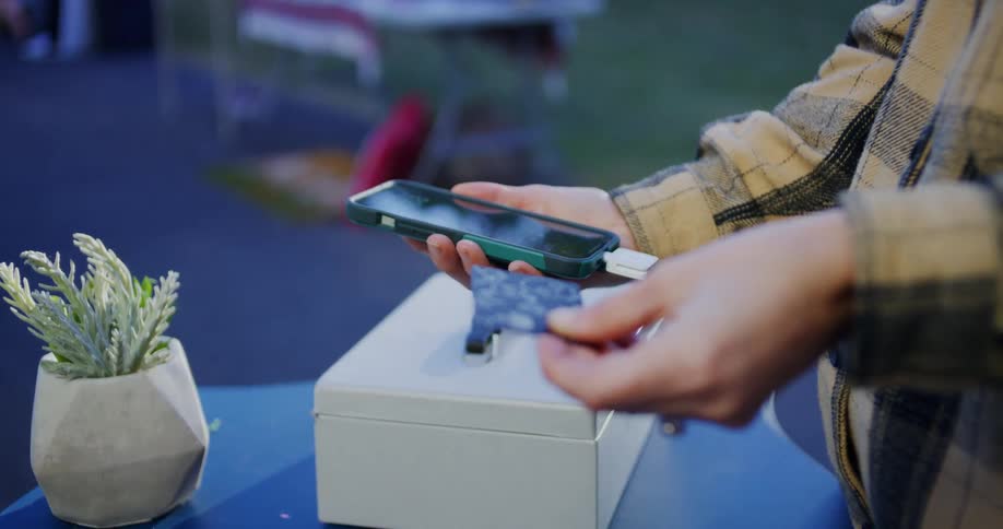 A close view shows a person in a plaid shirt swiping a credit card through a mobile reader attached to a smartphone to complete a purchase over a metal cash box on a blue table