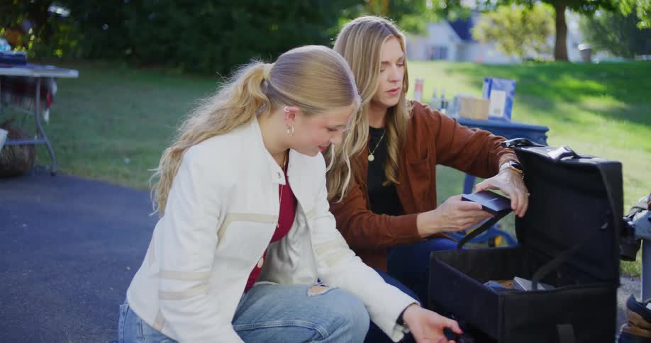 Two young women crouch to examine the contents of a black makeup case at an outdoor yard sale