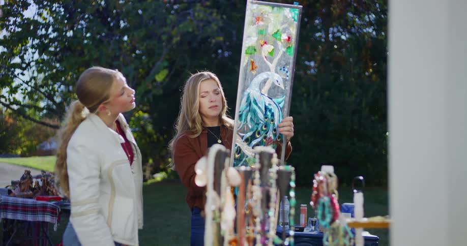 A woman wearing a brown sweater holds up a colorful glass artwork of a peacock for a young woman in a white jacket to view at an outdoor flea market on a sunny day
