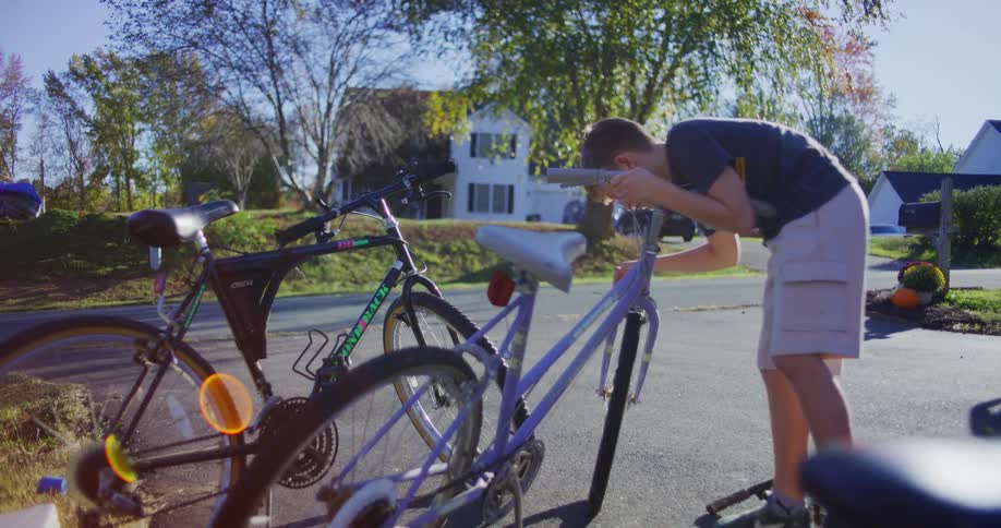 A young boy in a dark shirt and khaki shorts adjusts the handlebars of a purple bicycle next to two other bikes in a suburban driveway on a sunny day
