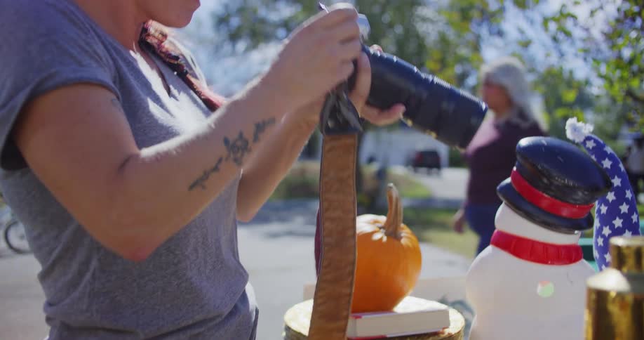 A close view of a woman with a braided hairstyle and tattoos holding and adjusting a large camera with a telephoto lens over a table of yard sale items on a sunny day