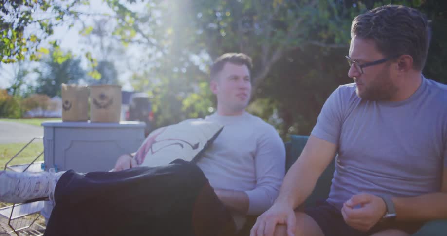 Two men sit outside on a couch on a sunny day talking with one man wearing a gray shirt and glasses and the other man in a white shirt holding a decorative pillow