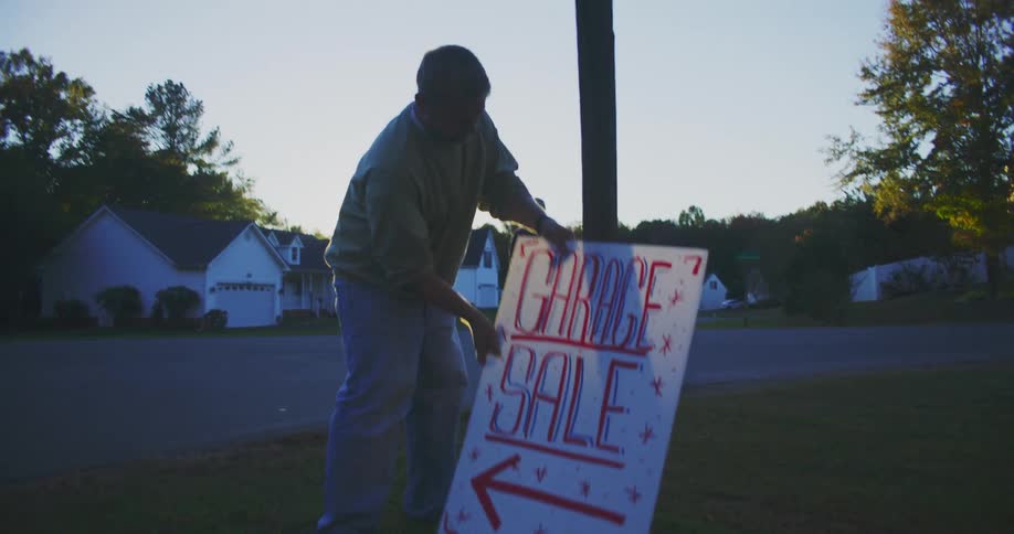 A man stands in a suburban street setting up a white sign that reads garage sale in red letters as the bright sun flares intensely from behind him
