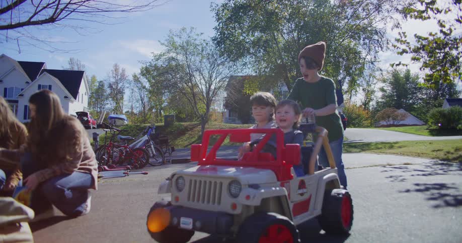 A young woman and three children interact with a ride on toy jeep in a suburban driveway filled with yard sale items on a bright sunny day