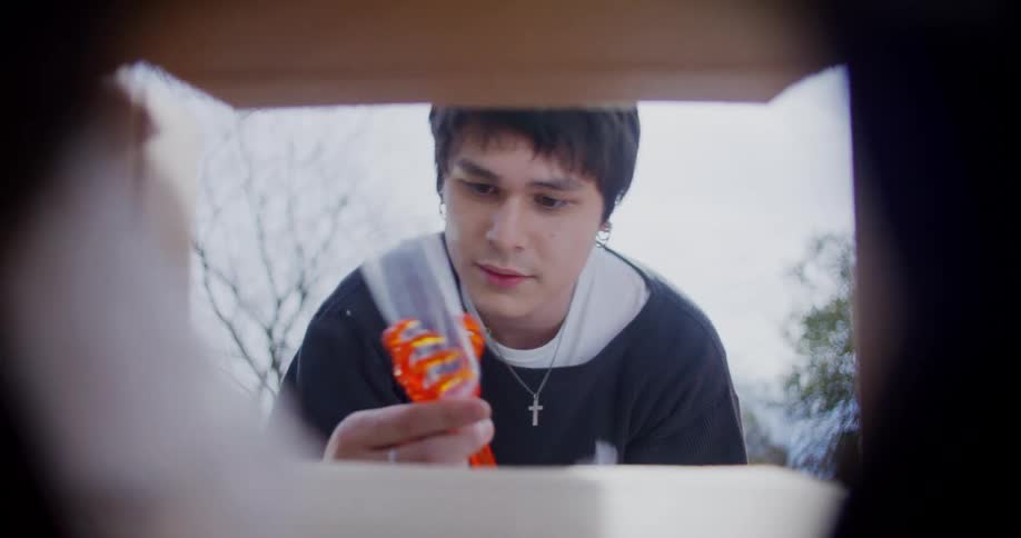 A teenage boy removes packaged goods from a cardboard box while standing outside