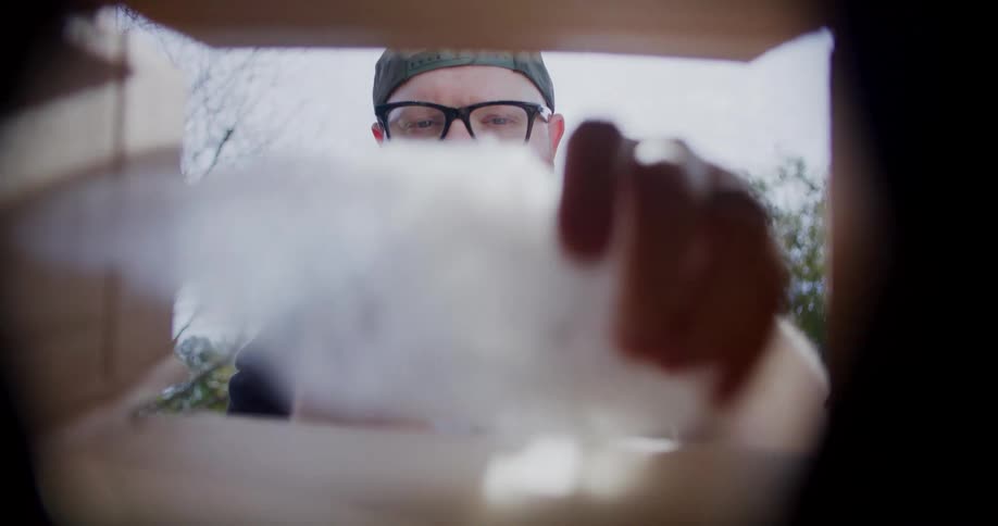 A point of view shot from inside an open package shows a bearded man wearing glasses and a cap removing bubble wrap and an item before turning and walking away outside on a bright day