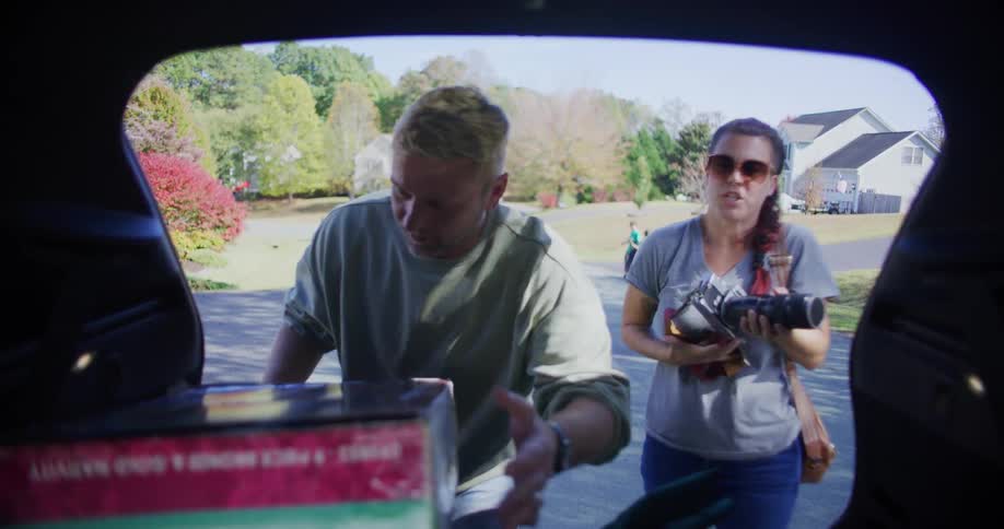 A man and a woman load a large cardboard box and a camera into the trunk of a car on a suburban street before waving goodbye to people walking away
