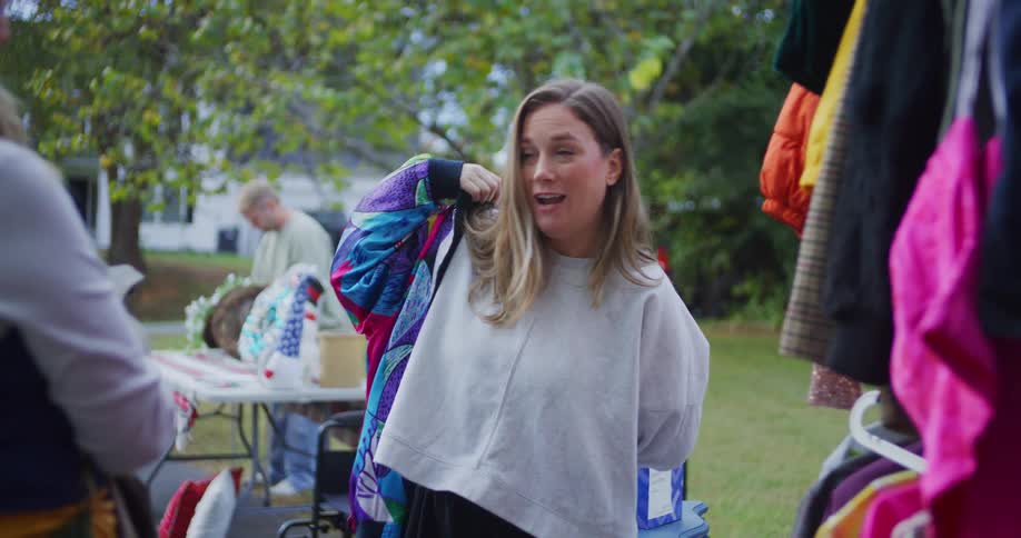 A woman is browsing a rack of clothes outdoors at what appears to be a yard sale or market and she enthusiastically pulls out a vibrantly patterned colorful vintage jacket she tries on the jacket over her grey sweatshirt checks the fit and smiles wit
