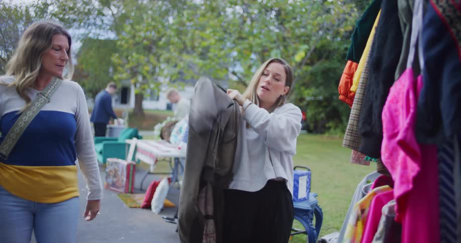 A woman in a light gray sweatshirt is looking through a rack of clothes at a yard sale she pulls out a brown leather jacket and tries it on looking for the zipperanother woman carrying a colorful bag approaches and interacts with her as they stand n