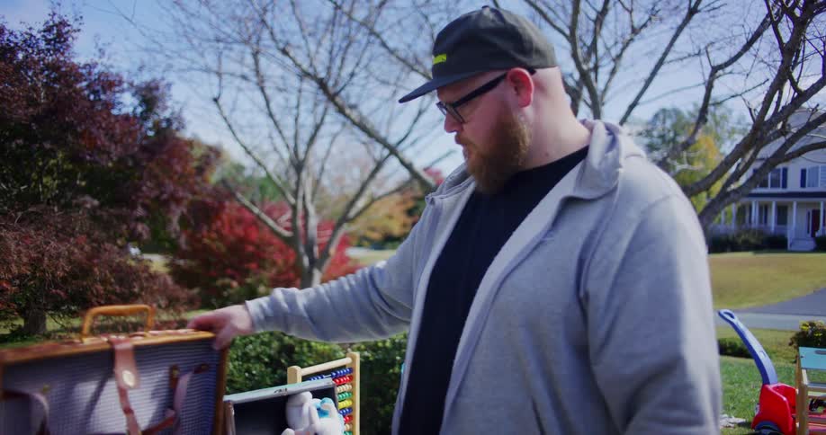 A bearded man in a gray hoodie opens a classic wicker picnic basket with a checkered interior to show a woman at an outdoor yard sale surrounded by colorful autumn foliage