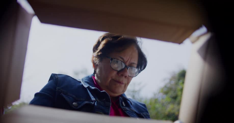 Woman wearing glasses looking inside a carboard box and removing an object wrapped in plastic outdoors during day time