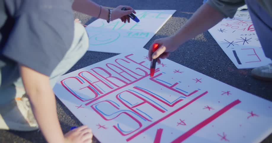 A closeup shot focuses on several pairs of hands kneeling on the ground actively drawing and filling in the large stylized red and blue letters for a garage sale sign on a white poster board