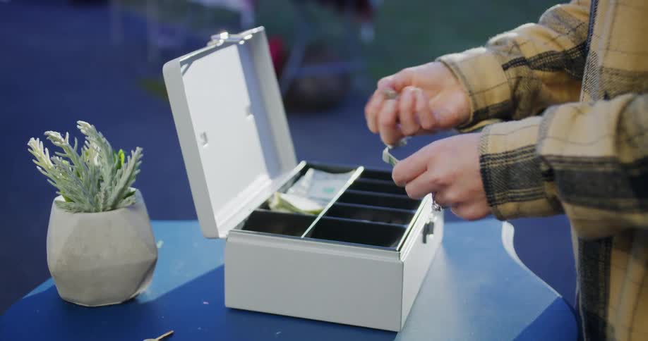 A close view shows a transaction taking place at an outdoor market as a person takes cash out of an open gray cash box and makes change for a customer