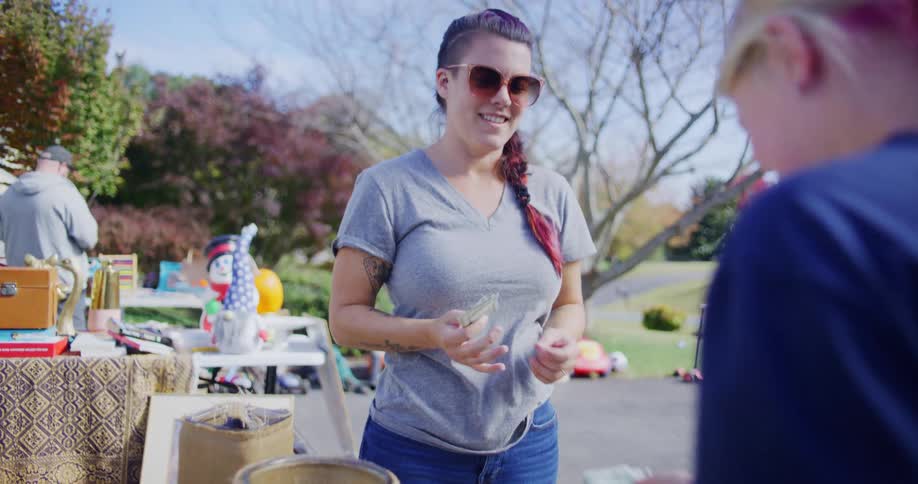 A woman with sunglasses and a gray shirt holds a camera and counts money next to a cash box and various items for sale at a sunny outdoor yard sale