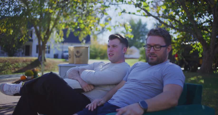 Two men are sitting closely together on a green couch outdoors on a sunny day possibly taking a break during a yard sale or moving event they transition from having their arms crossed and appearing relaxed or bored to beginning a conversation with each