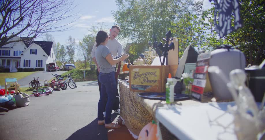 A man and a woman stand by a table piled high with garage sale items as the woman takes a picture of the merchandise with a camera on a bright sunny day