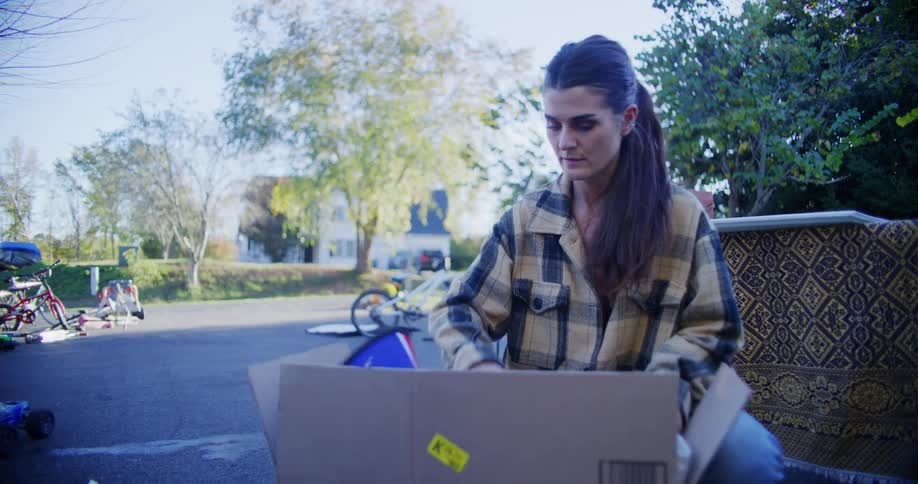 A young woman packs items into a cardboard box then stands and lifts it behind her a young man picks up a desk both working together in their driveway