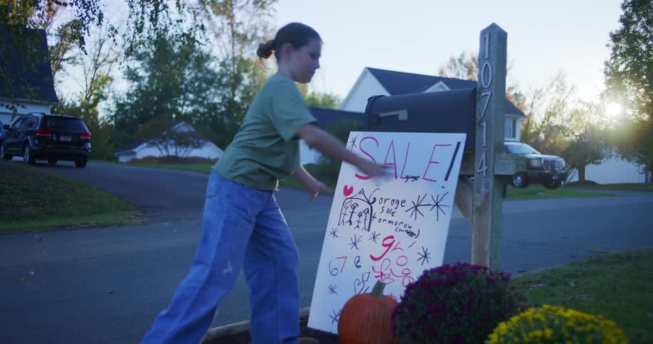 A girl leans a homemade garage sale sign against a mailbox post
