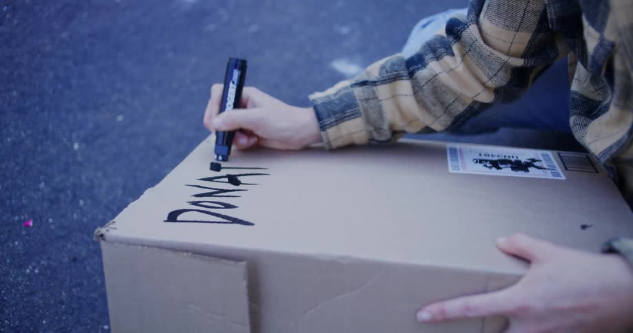 A closeup shot shows a persons hands holding down a cardboard box and using a black marker to write the word donations in large stylized letters on the top flap