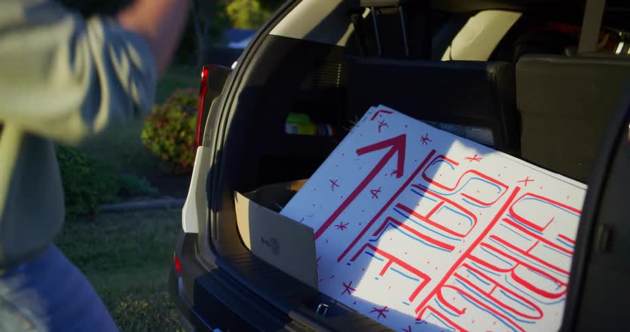 A man places a sign in the trunk of his car then closes it and gets into the vehicle under the bright sun of a clear day