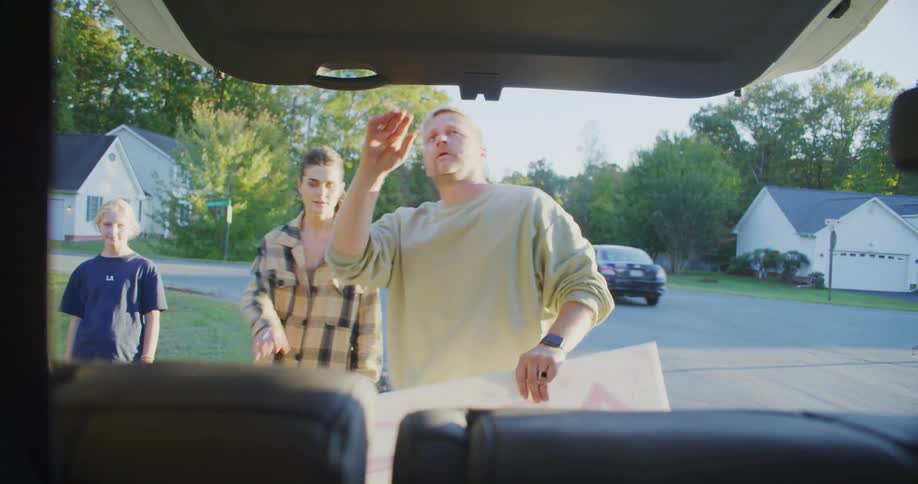 Home retrieves a large poster from the trunk of a car while two others watch in the background