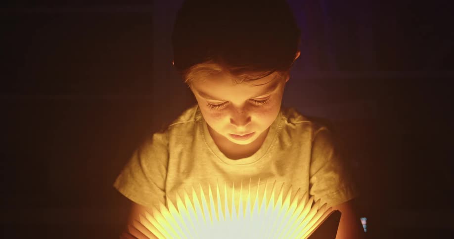 A close view of a young boy in a dimly lit room looking intently down at a glowing light source from a fanlike paper book or light object that illuminates his face