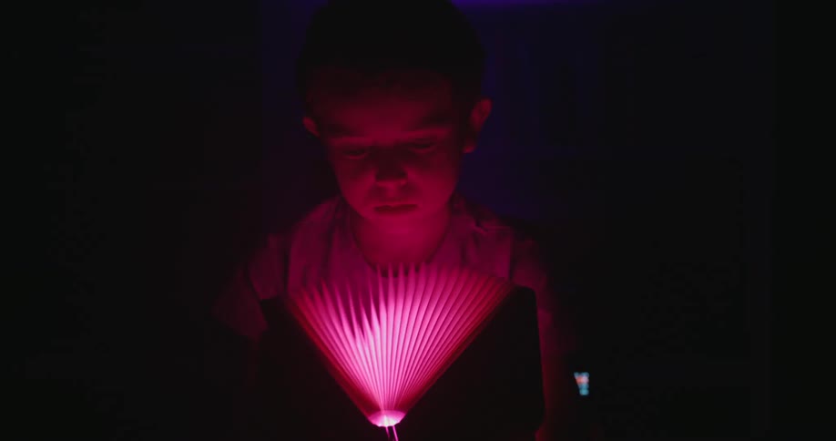 A close view of a young boy in a dark room looking down at a fanlike paper light object that changes colors from warm yellow to cool blue and red illuminating his face