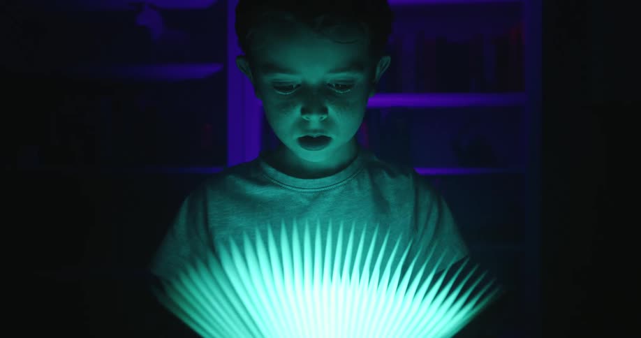 A young boy opens a turquoise glowing book in a dark room