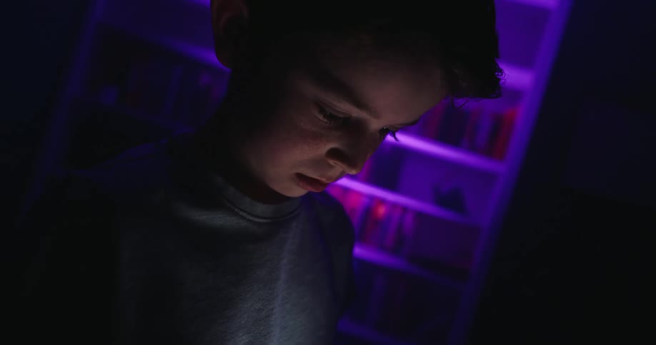 The video shows a closeup of a young boy in a dark room illuminated only by the light from a smartphone he is holding and looking down at a bookshelf behind him is cast in a vibrant purplepink light